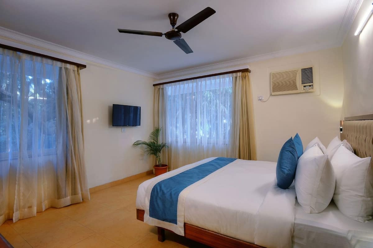 Hotelkamer met een bed, gordijnen, een plafondventilator, een televisie, een potplant en een radiator.