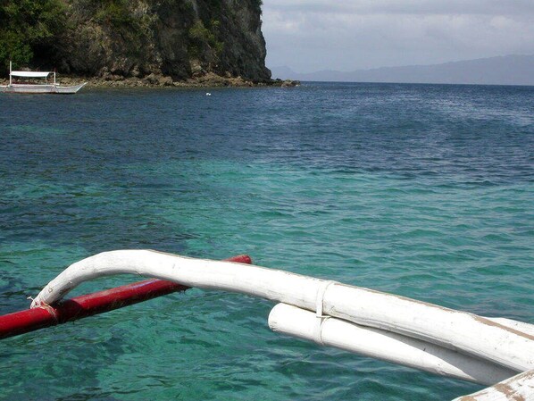 A boat with a red and white structure, clear turquoise water, and a rocky cliff in the background. - Out of the Blue Resort (Puerto Galera)