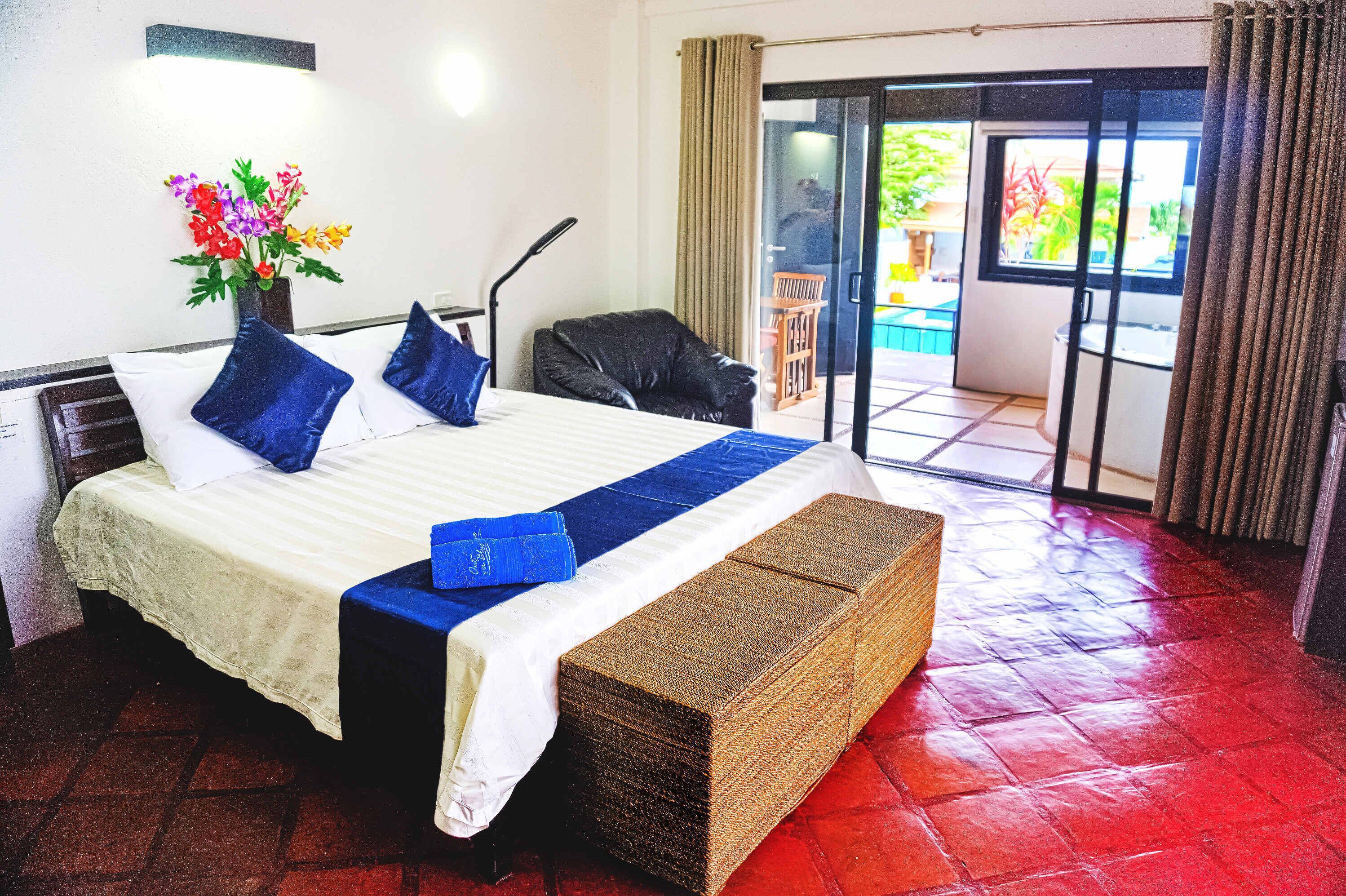 A hotel room with a bed, a chair, a small table, a vase with flowers, and a view of the outside.