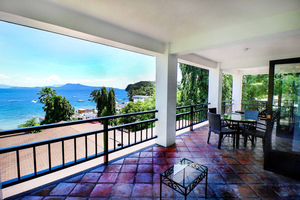 Een balkon met uitzicht op zee en bergen, met een tafel en stoelen.