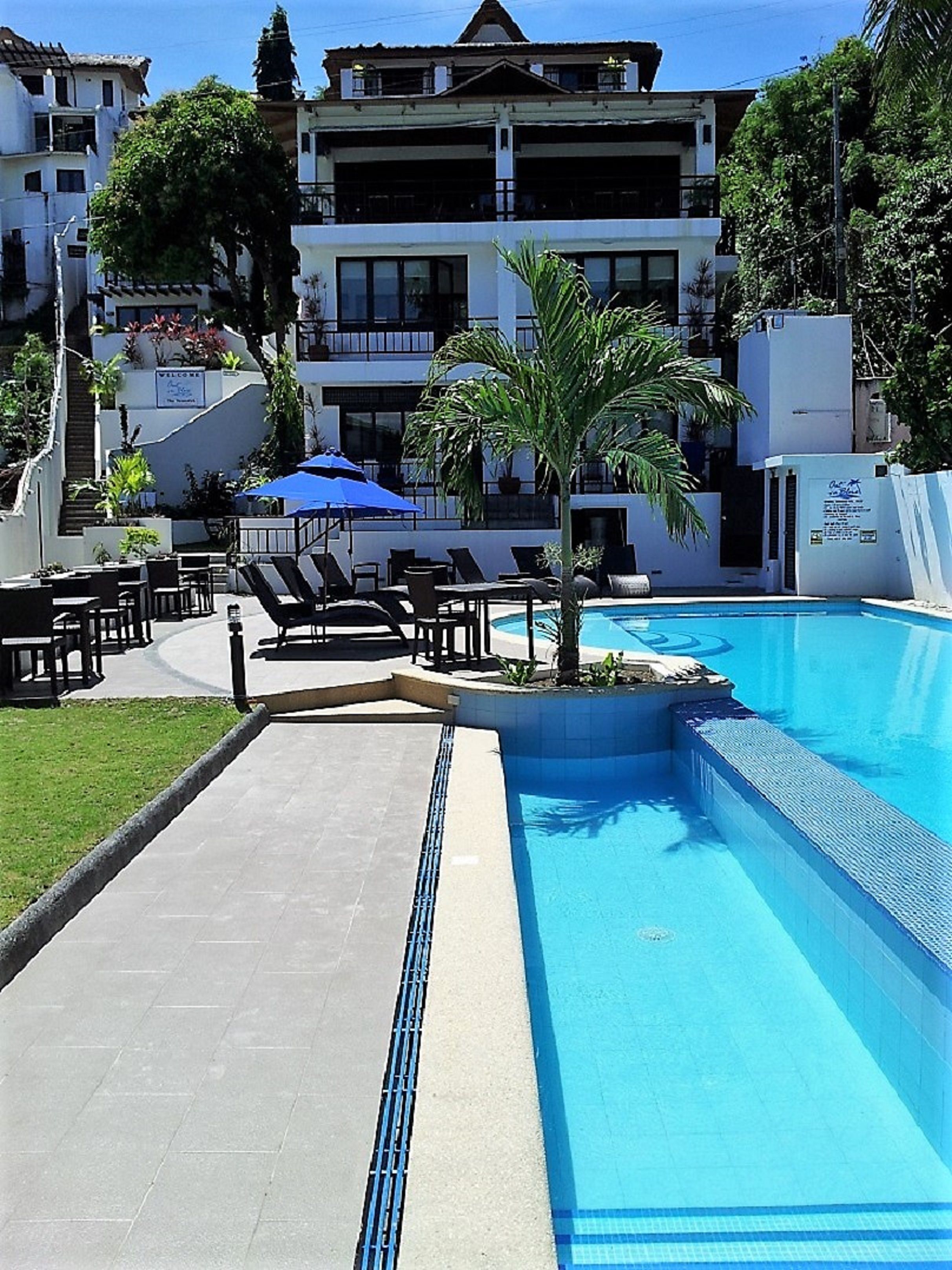 Una piscina con sillas de descanso y sombrillas, rodeada de edificios y vegetación.