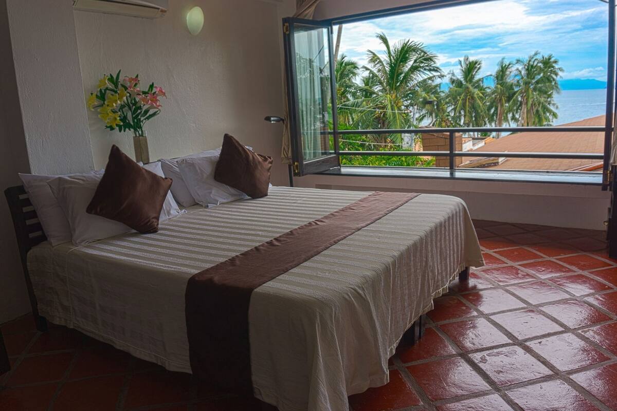 Een hotelkamer met een bed, een raam met uitzicht op palmbomen en de zee, en een bloemenvaas aan de muur.