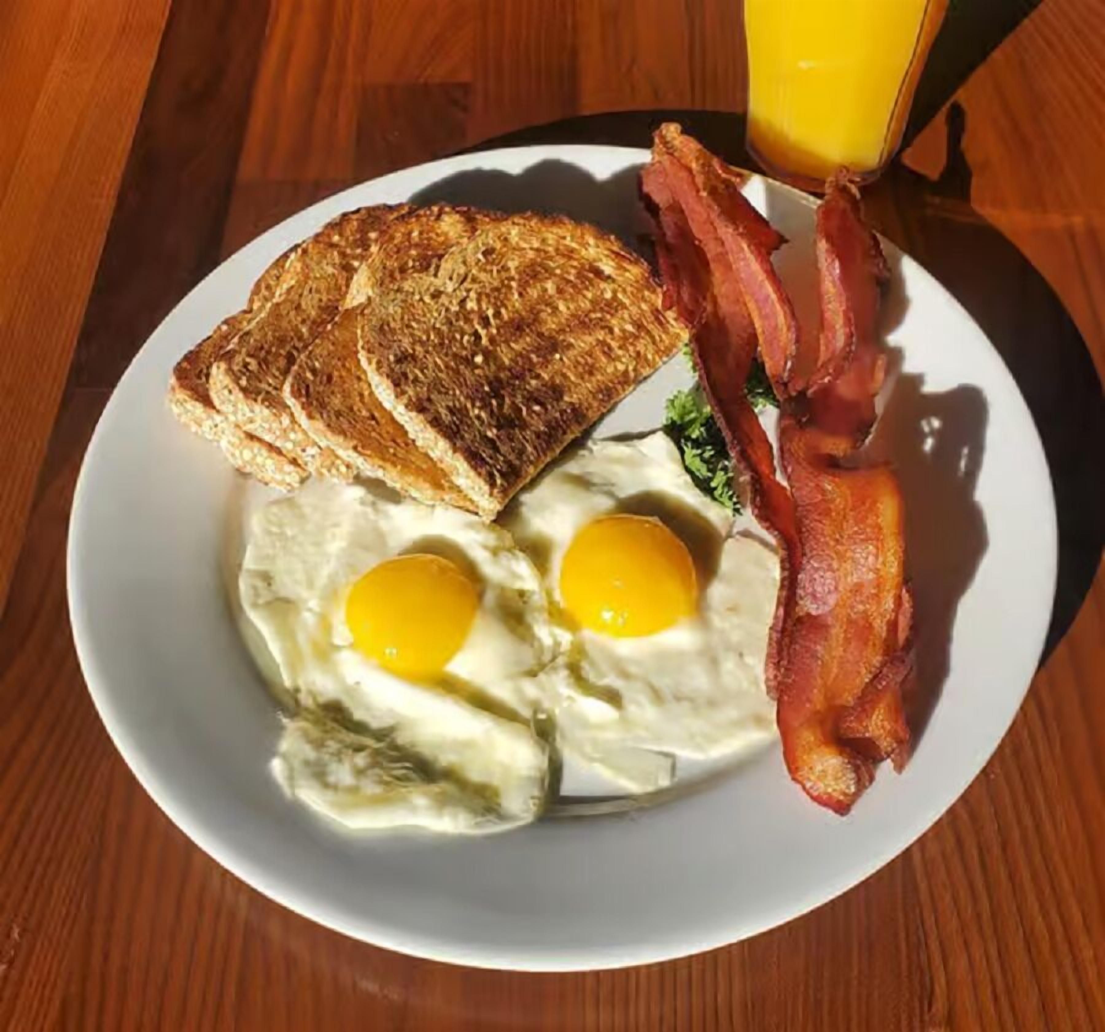 Un plato con dos huevos fritos, tostadas, tocino y un vaso de jugo de naranja.