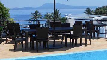 A poolside area with a table and chairs under a blue umbrella, overlooking a body of water and distant mountains.