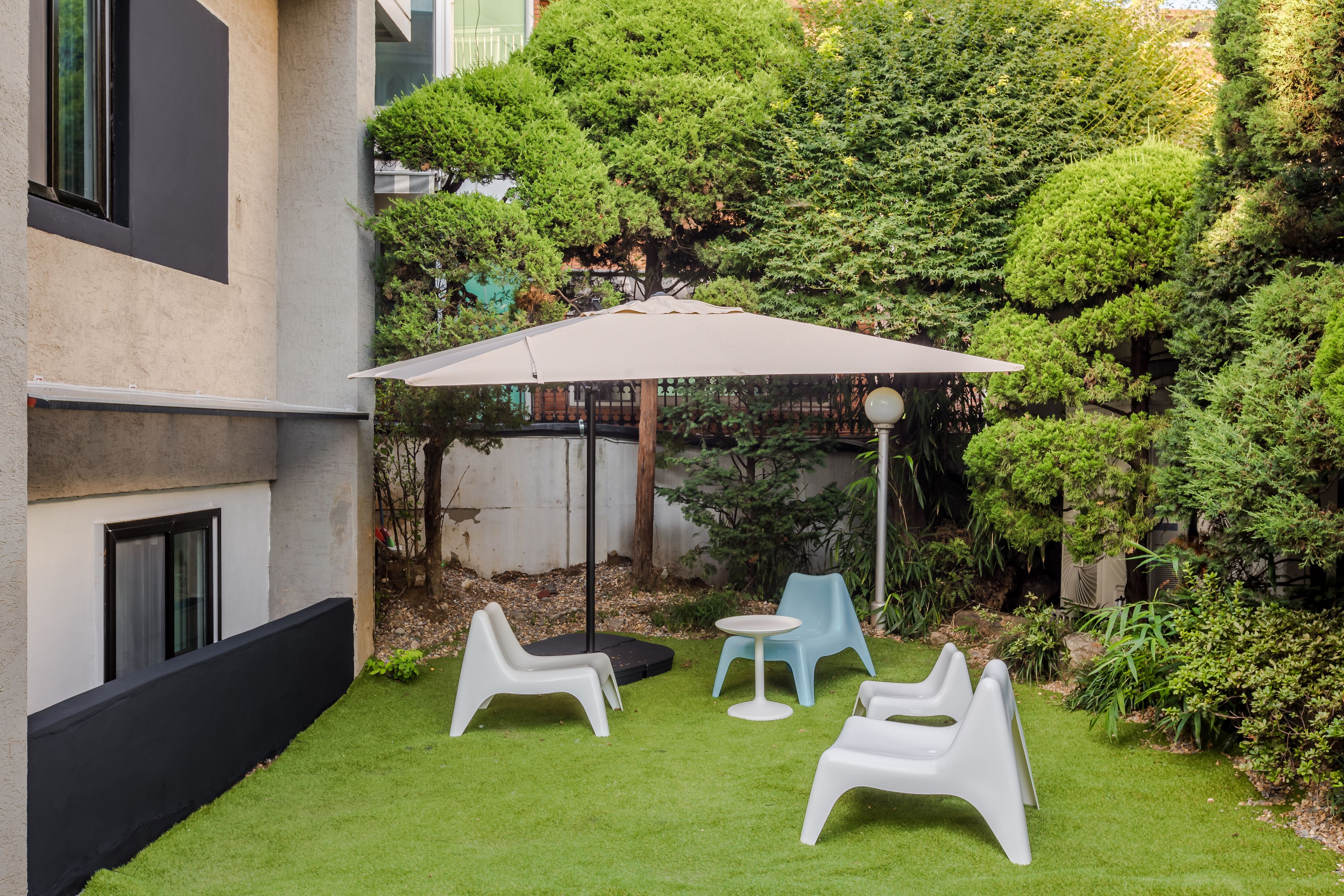 An outdoor seating area with white plastic chairs and a table, surrounded by lush greenery and a large umbrella.