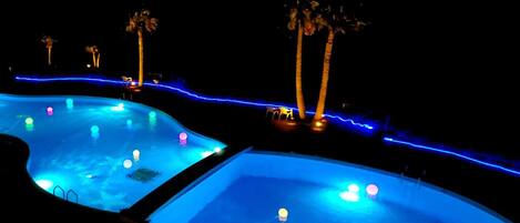 Vue nocturne d’un espace piscine comprenant deux piscines, des palmiers et un éclairage d’ambiance bleuté.