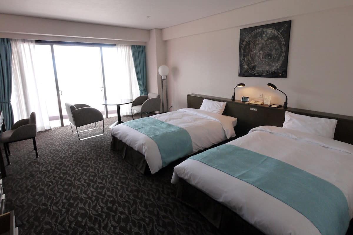 Quarto de hotel com duas camas, uma escrivaninha, uma cadeira e uma janela grande com cortinas.