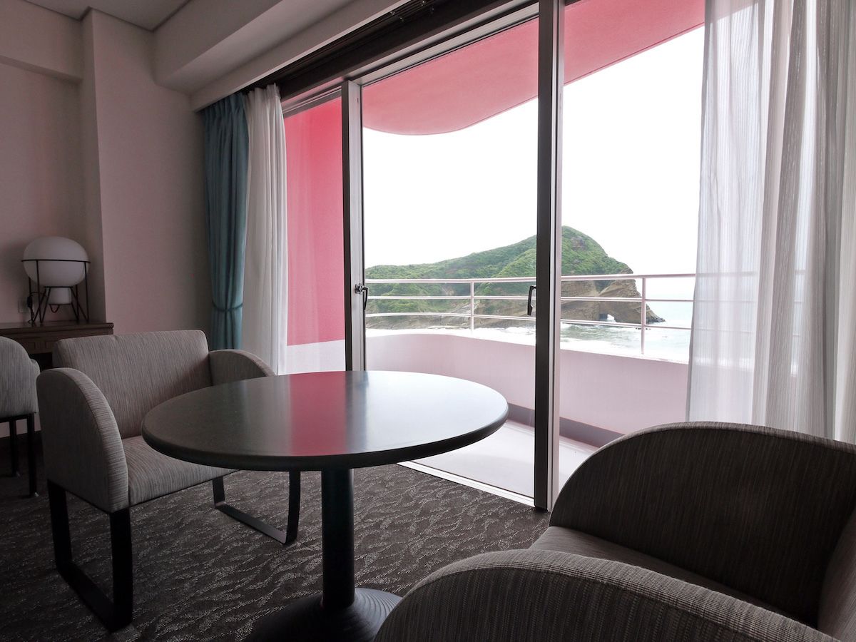 A hotel room with a balcony, a round table, two armchairs, and a view of a mountainous landscape.