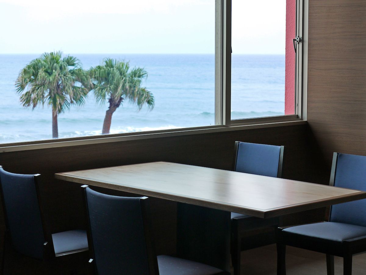 A dining area with a table and chairs, a window with a view of the ocean and palm trees.