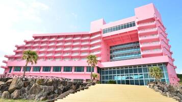 A pink multi-story hotel with a central glass facade, a staircase leading to the entrance, and palm trees in front.