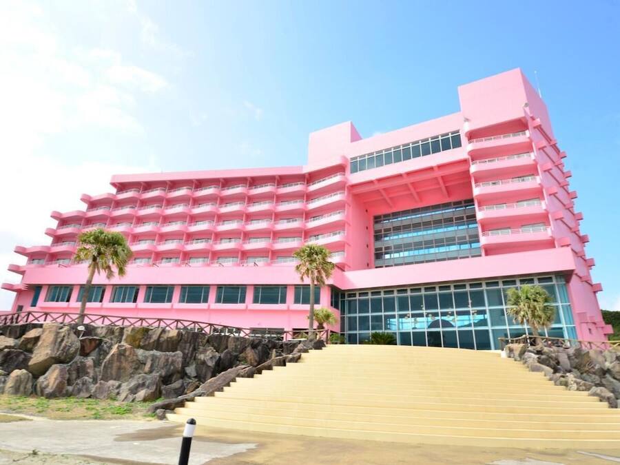 A pink multi-story hotel with a central glass facade, a staircase leading to the entrance, and palm trees in front.
