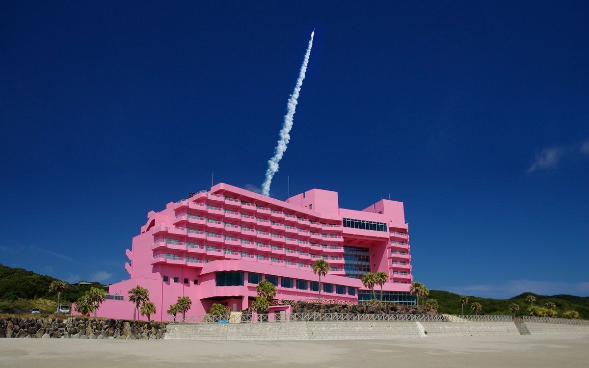 A pink hotel with multiple floors, a balcony on the second floor, and a clear sky.