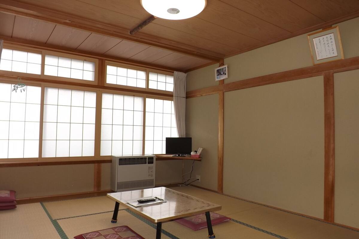 A traditional Japanese room with tatami flooring, a tatami mat in the center, a small table with a remote control, a heater, and a window with a view.