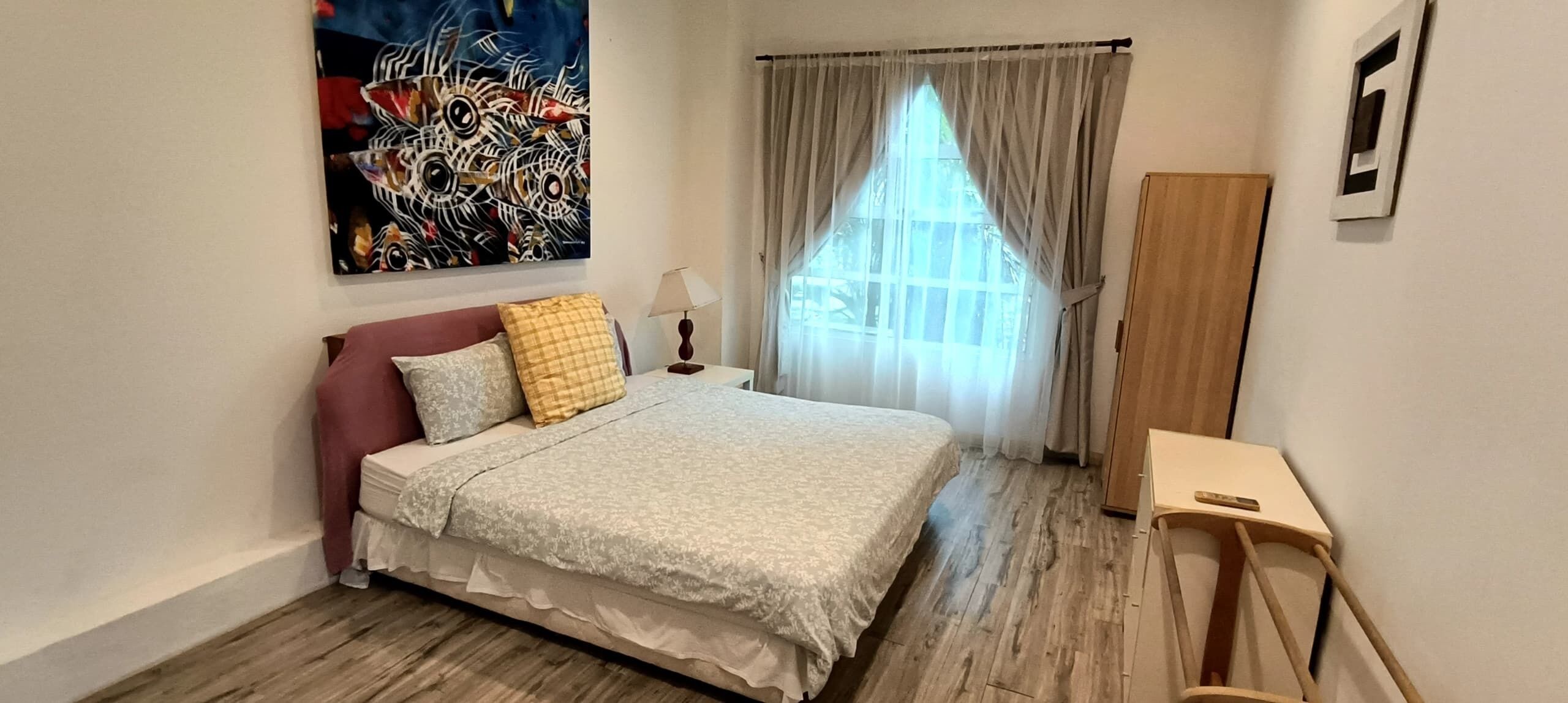 A bedroom with a bed, a bedside table, a lamp, a window with curtains, and a painting on the wall.