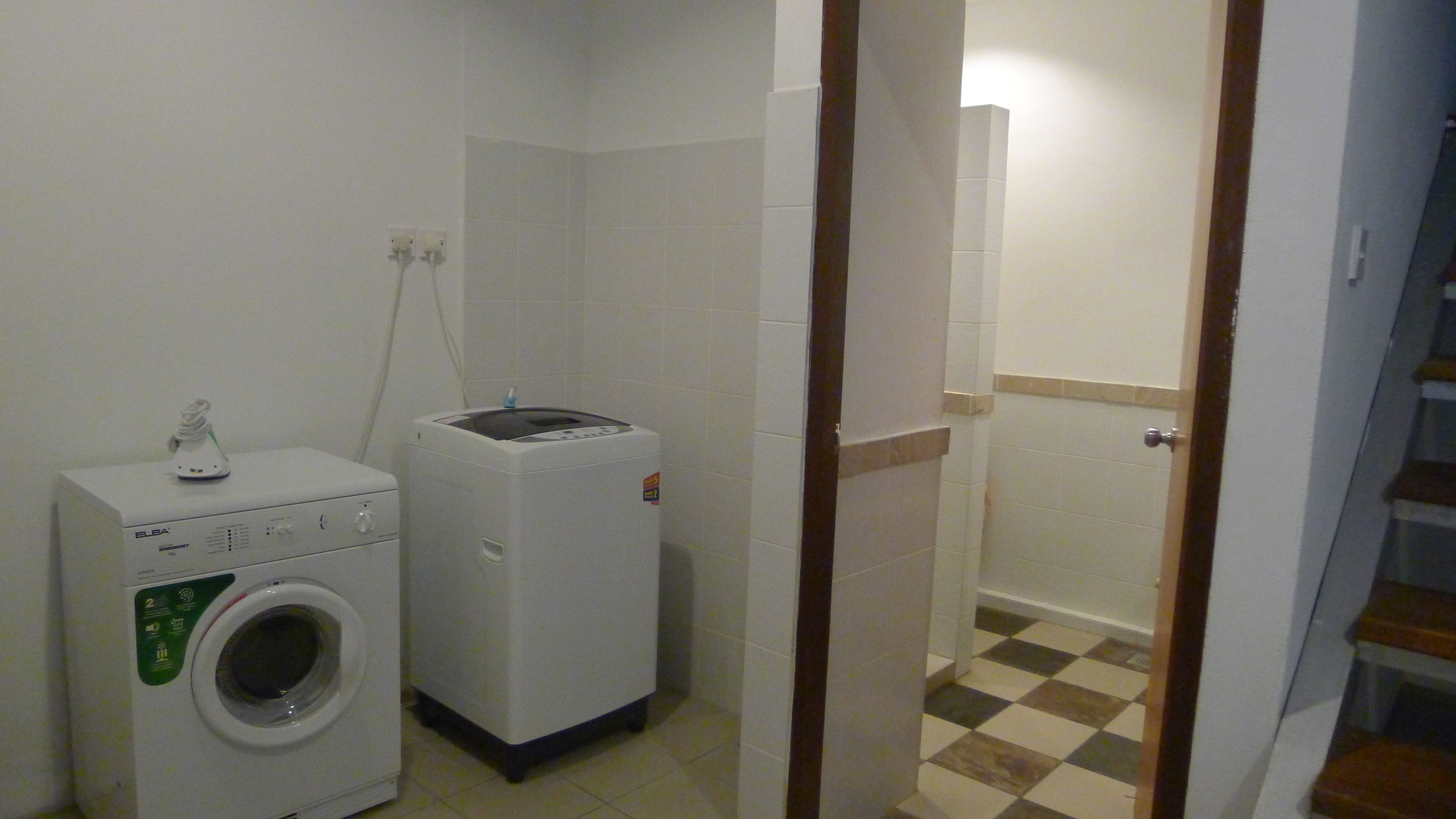 A laundry room with a front-loading washer and dryer, a small table with a lamp, and a door leading to another room.