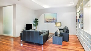 Una sala de estar moderna con un sofá, una mesa de centro y un televisor. Se observa una estantería a la derecha y un cuadro en la pared.