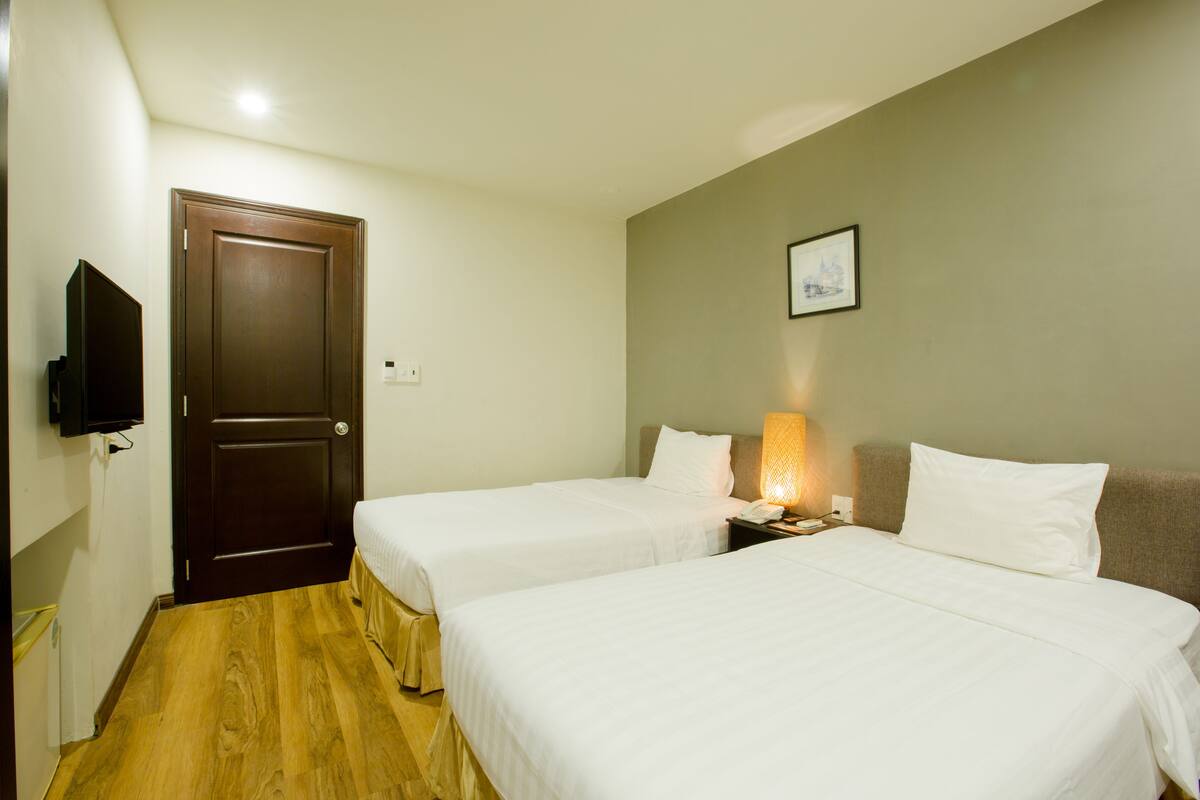 A hotel room with two beds, a wooden floor, a TV mounted on the wall, and a framed picture on the wall.