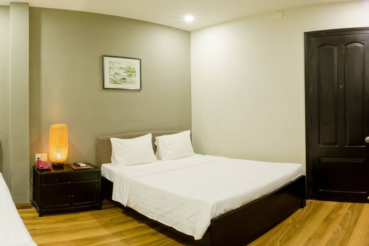 A hotel room with two single beds, a nightstand with a lamp, a framed picture on the wall, and a dark door.