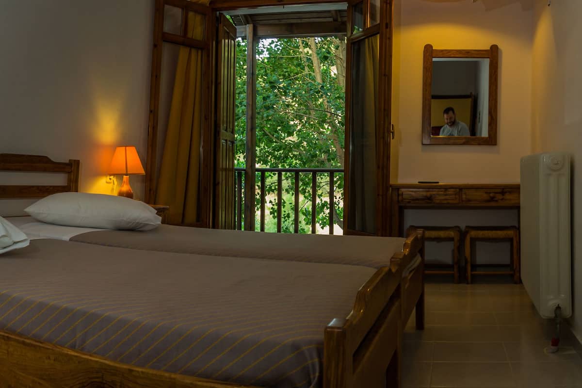 A double bed with a wooden headboard, two bedside lamps, a small table with a lamp, a mirror, and a window with a view of trees.