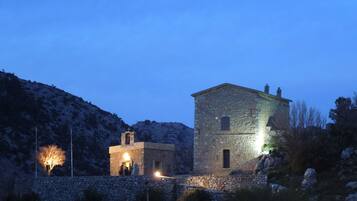 Un edificio de piedra con un campanario, rodeado de terreno rocoso y vegetación escasa.