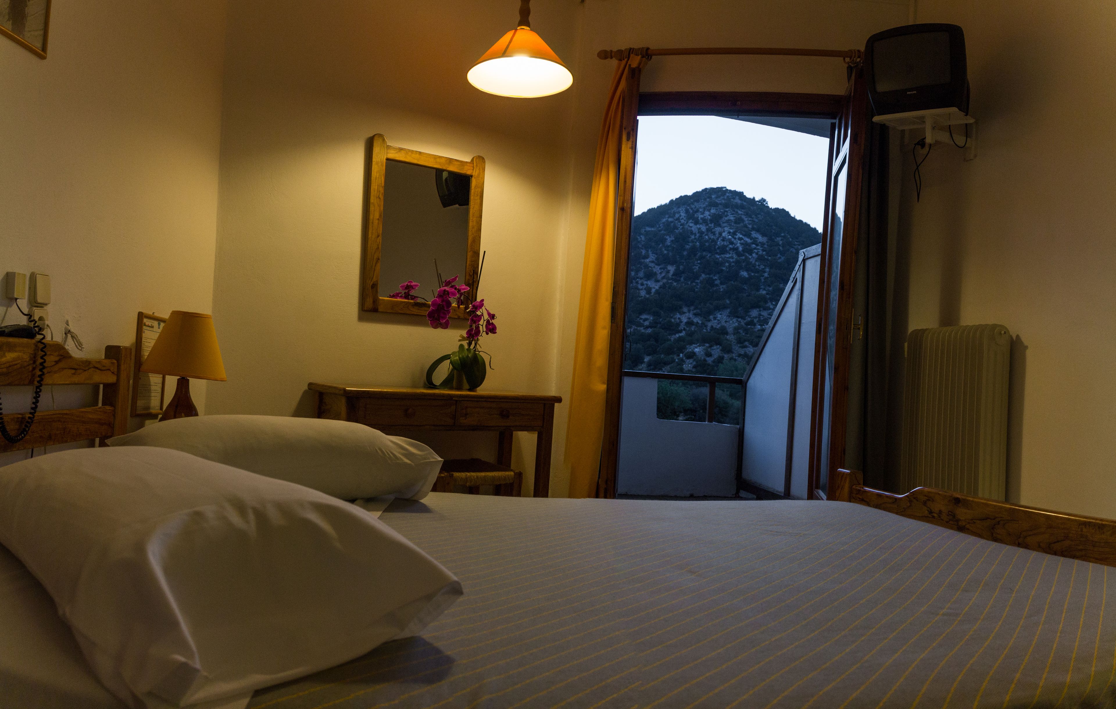 Quarto de hotel com uma cama, uma escrivaninha, um espelho, um abajur e vista para uma montanha.