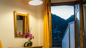Une chambre à coucher avec un lit, une table de chevet ornée d’un vase de fleurs, un miroir et une vue sur un balcon avec une montagne en arrière-plan.