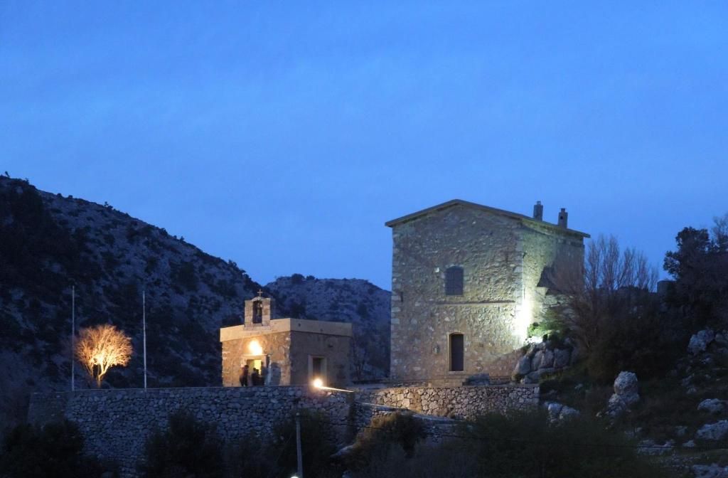 Un edificio de piedra con una pequeña torre de campanas, rodeado de terreno rocoso y vegetación escasa.