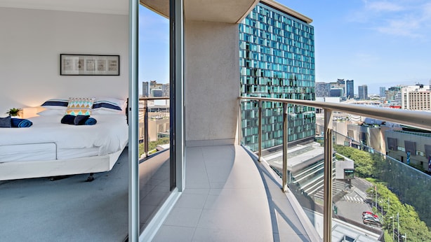 Een moderne slaapkamer met een groot bed, stadszicht en een balkon met glazen hekwerk.