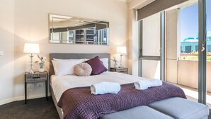 Een hotelkamer met een groot bed, twee nachtkastjes met lampen, een groot raam met uitzicht op gebouwen, en een grijze bank.