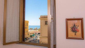 Una habitación con un amplio ventanal que da a una calle y edificios, una obra de arte enmarcada en la pared y una cortina entreabierta.