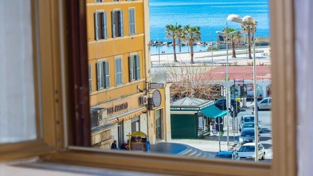 Vista desde una ventana a una calle con un edificio, coches aparcados y una playa con palmeras.