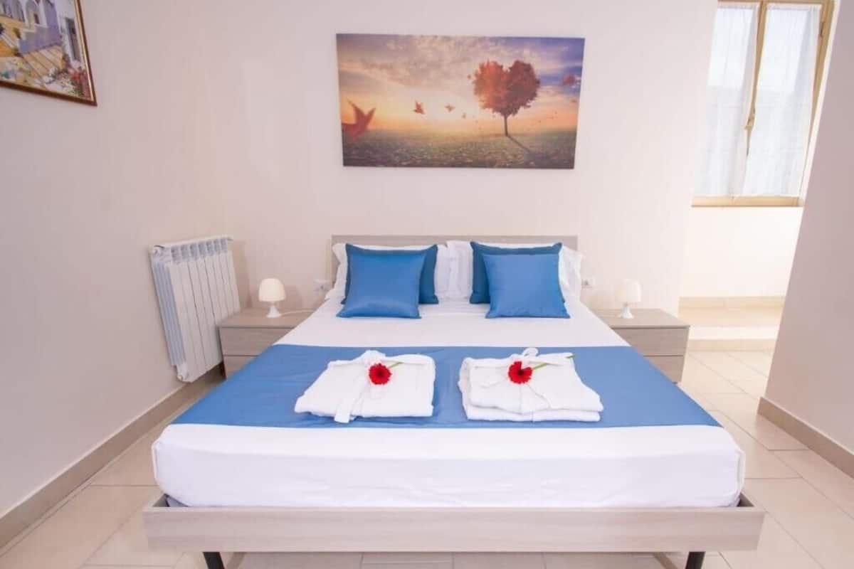 Ein Hotelzimmer mit einem großen Bett, blauer und weißer Bettwäsche, zwei Handtüchern mit roten Blumen, einem Bild an der Wand und einem Fenster mit Vorhängen.