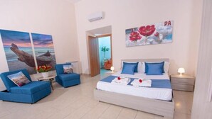 Un dormitorio con una cama, dos sillones azules, una mesita con un florero y un cuadro de una puesta de sol.