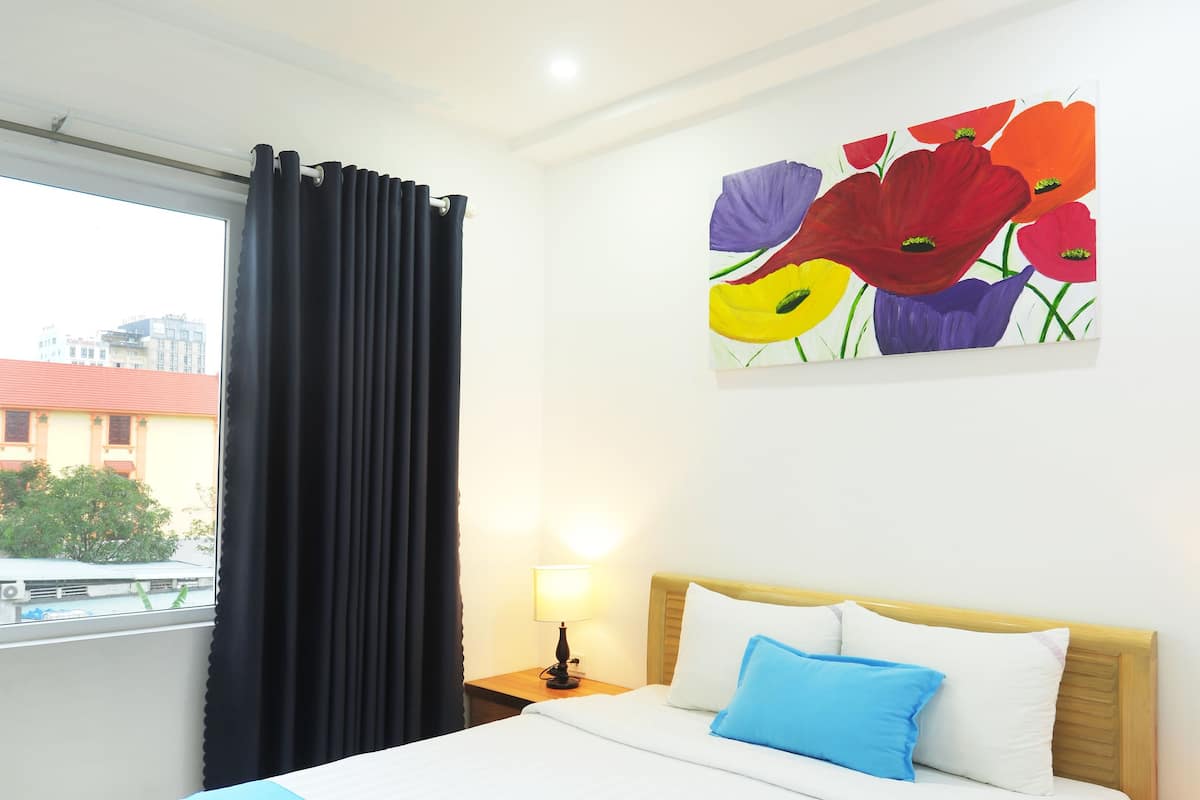 Een hotelkamer met een bed, een nachtkastje, een lamp, een raam met gordijnen en een schilderij aan de muur.