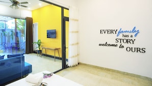 A hotel room with a yellow wall, a ceiling fan, a bed with a white comforter, a small table with a plant, and a wall sign welcoming guests.