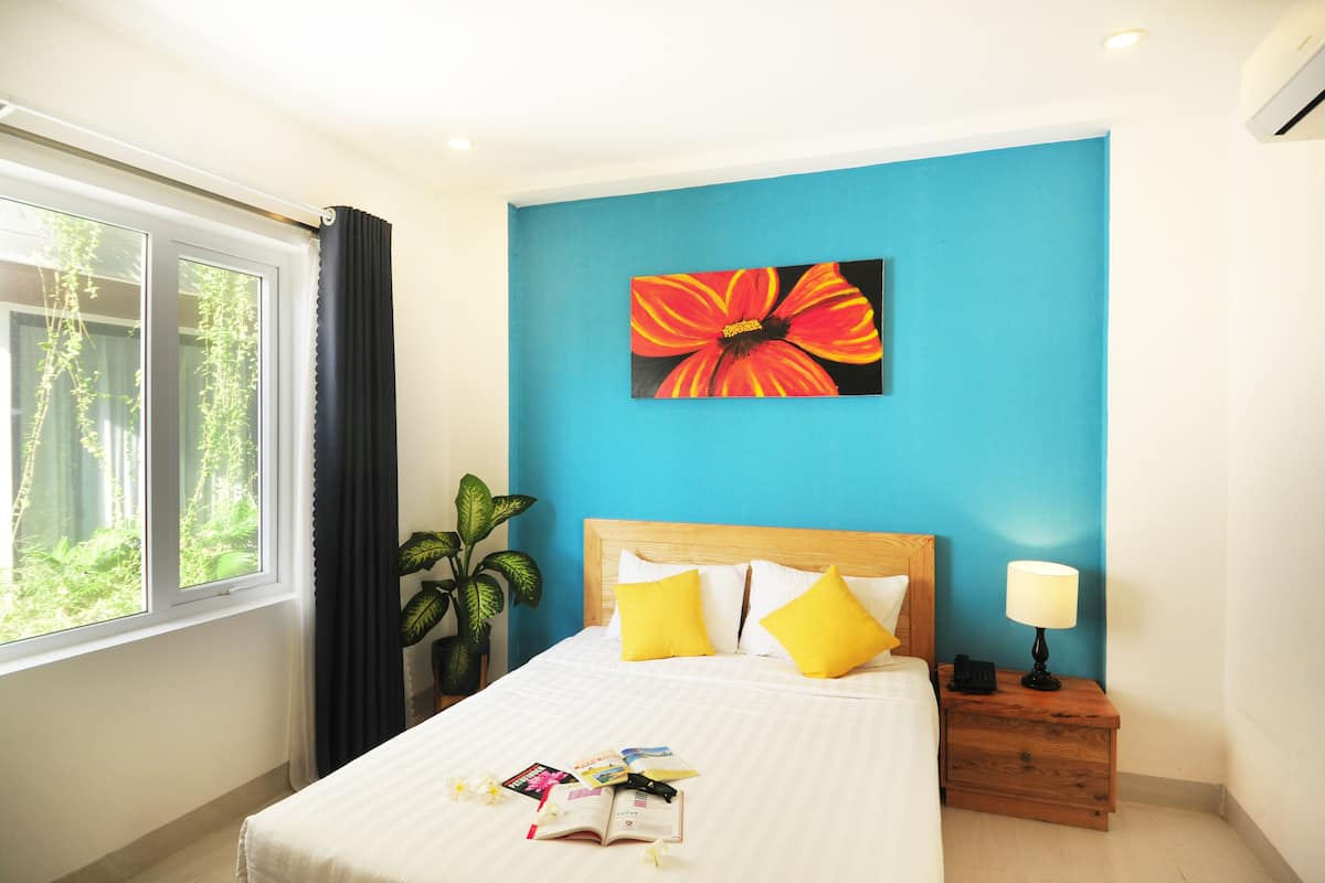 Een hotelkamer met een bed, nachtkastje, lamp, raam met gordijnen en een bloempot met plant.