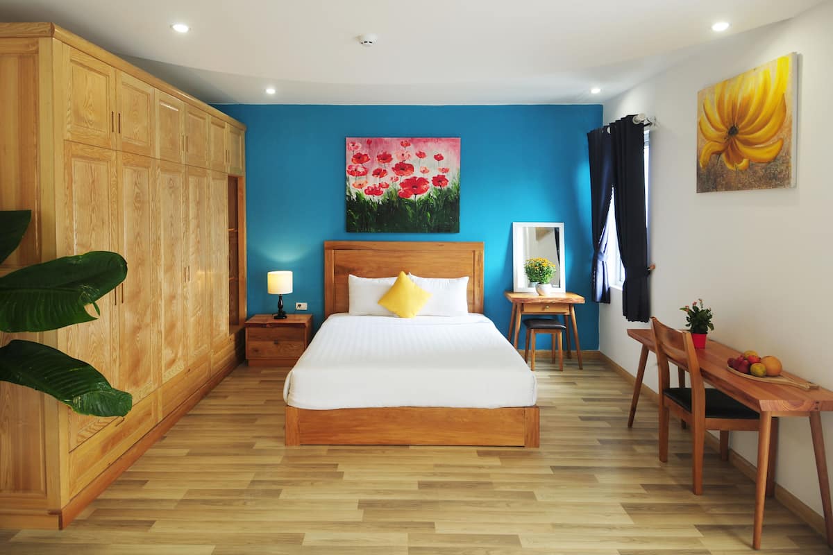 Een slaapkamer met een houten garderobe, een bed met wit beddengoed, een bureau met stoel, en een bloemstillevoering aan de muur.