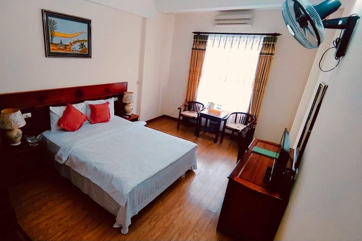 A hotel room with a large bed, a desk, two chairs, a fan, and a painting on the wall.