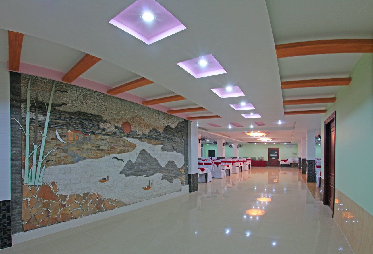 A hotel lobby with a long corridor, a mural on the left wall, and a row of tables with chairs on the right.