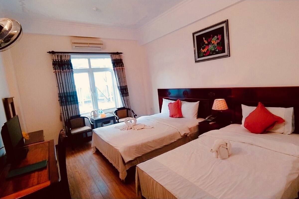 A hotel room with two beds, a desk, a chair, a window with curtains, and a ceiling fan.