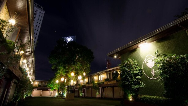 A courtyard with a large tree, a building with a sign, and a view of a cityscape at night. - Victoria Court Canley - Pasig (Pasig)