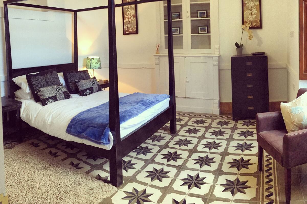 Ein Bett mit Baldachin, mit blauer Decke, einem gemusterten Teppich, einer Kommode und einem Sessel in einem Raum mit Sternenmuster auf dem Boden.