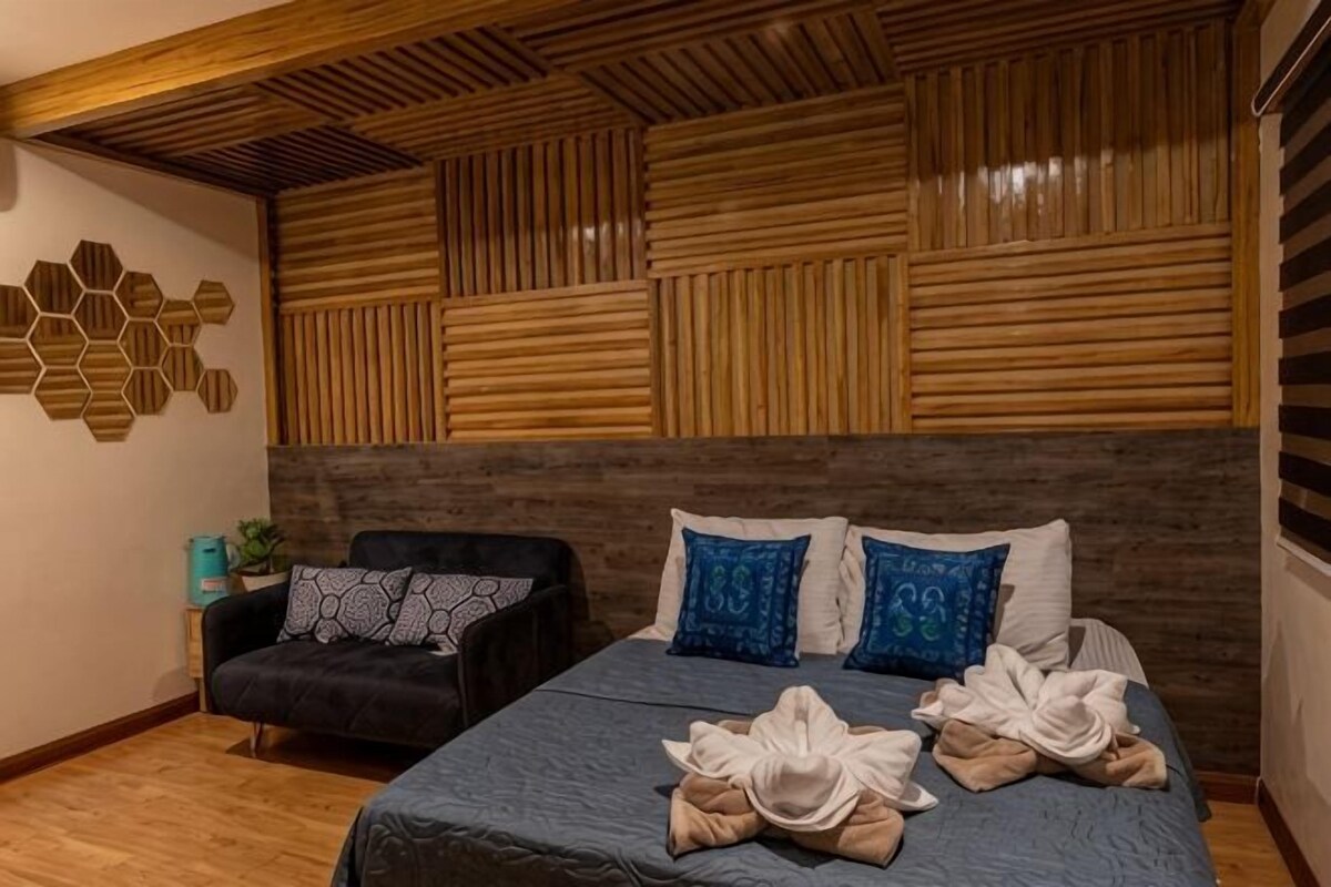A hotel room with a wooden headboard, a bed with multiple pillows, a black armchair, and a wooden floor.
