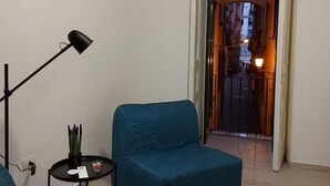 Un salon avec un canapé bleu, une petite table d’appoint, un lampadaire et une vue sur la rue, visible à travers une porte ouverte.