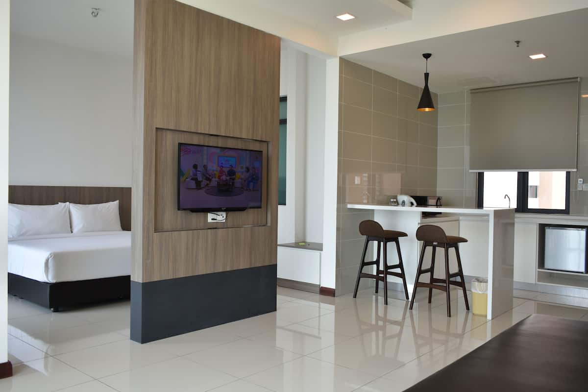 A modern hotel room with a bed, a TV mounted on a wooden panel, a kitchen area with a bar stool, and a small island.