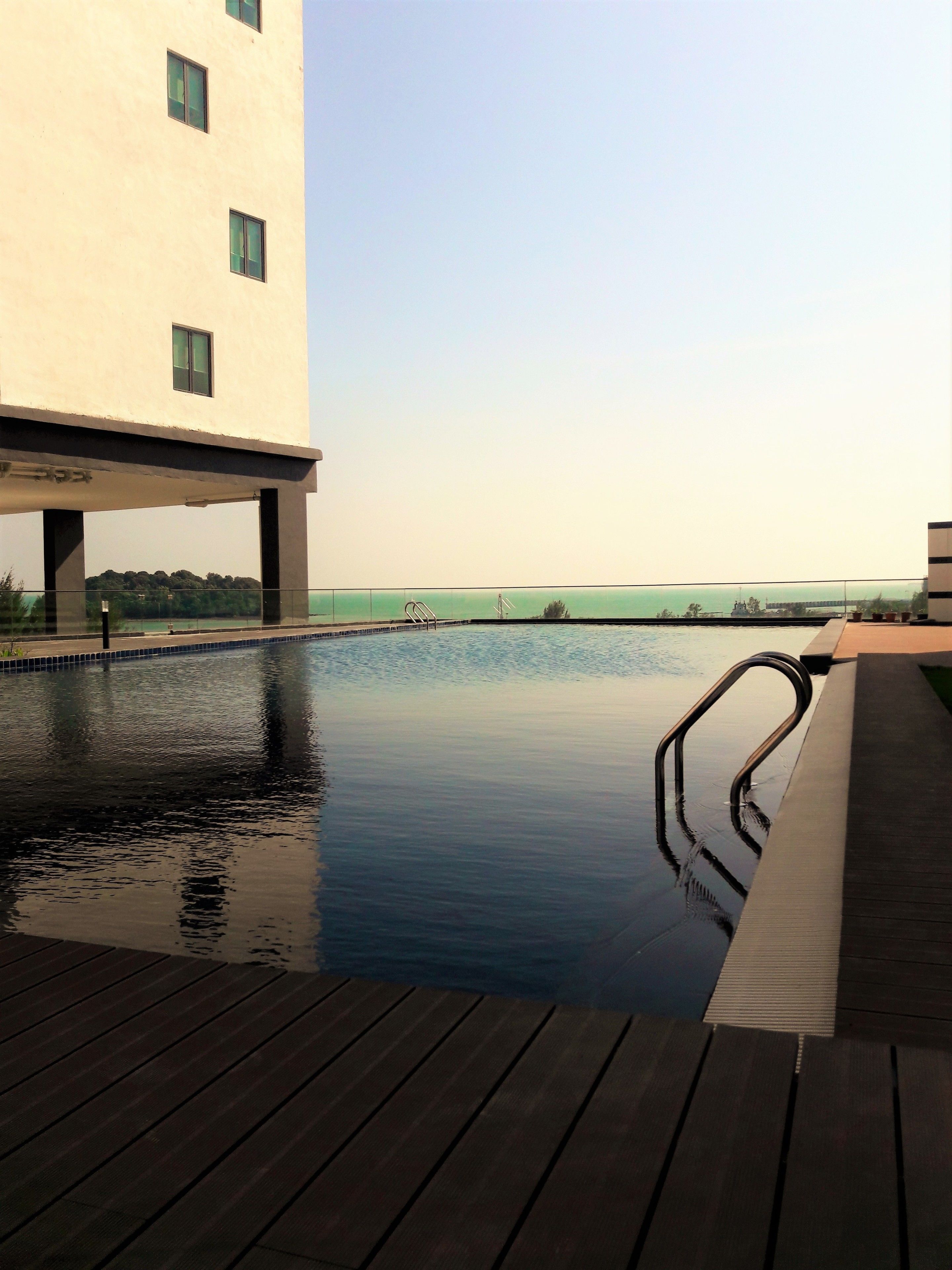 A swimming pool with a wooden deck, a view of the sea, and a building in the background.