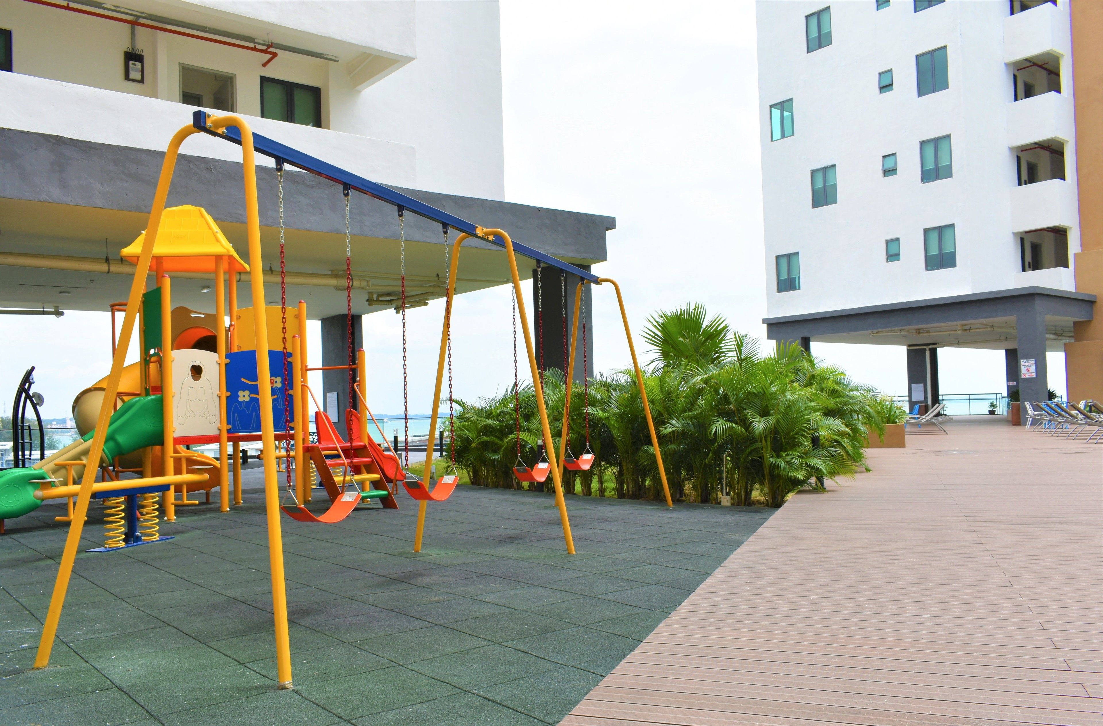 Un patio de juegos al aire libre con columpios y toboganes, rodeado por un edificio con muchas ventanas y una palmera.