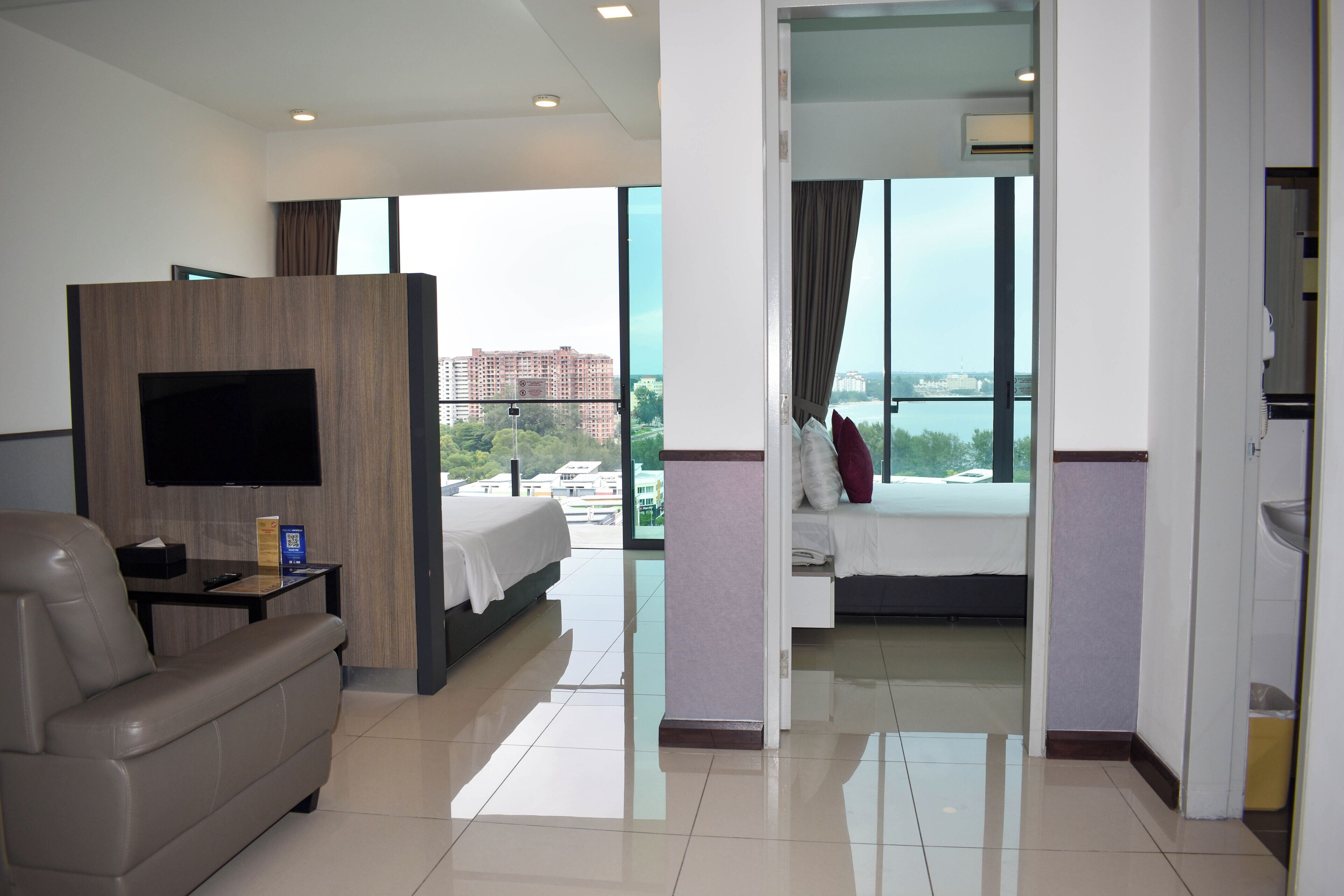 A hotel room with a large bed, a TV mounted on the wall, a sofa, and a view of the city through large windows.
