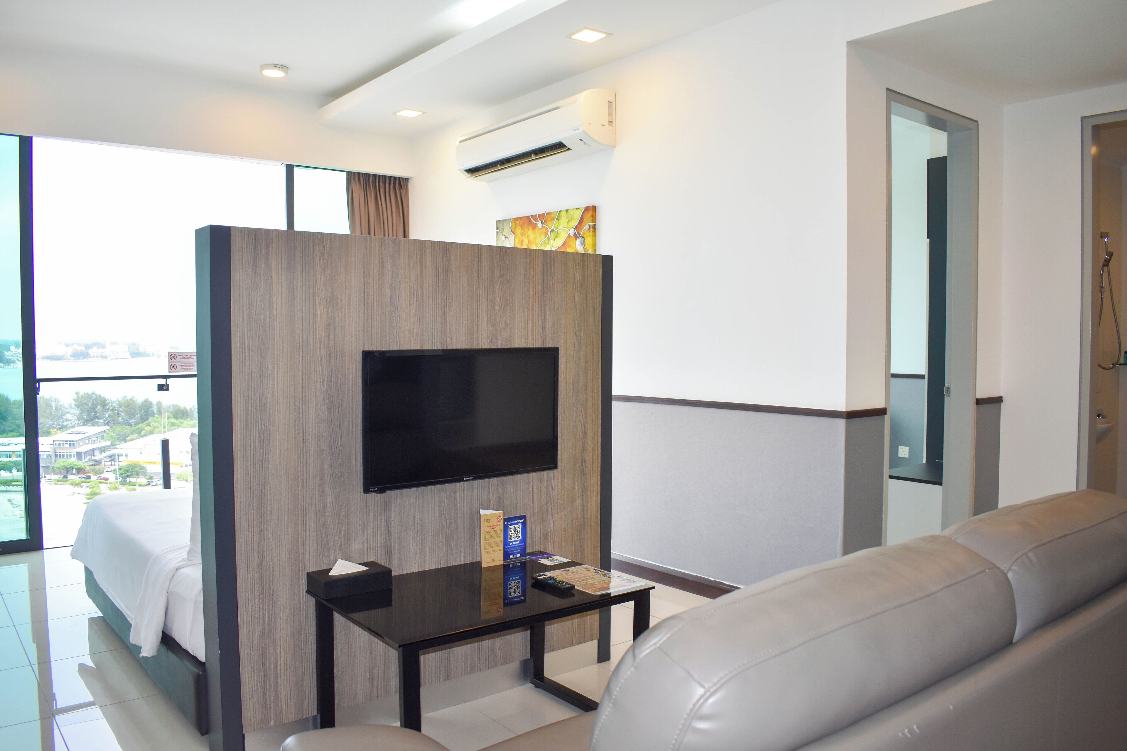A modern hotel room with a bed, a TV mounted on a wooden partition, a leather sofa, and a small table with a remote control.