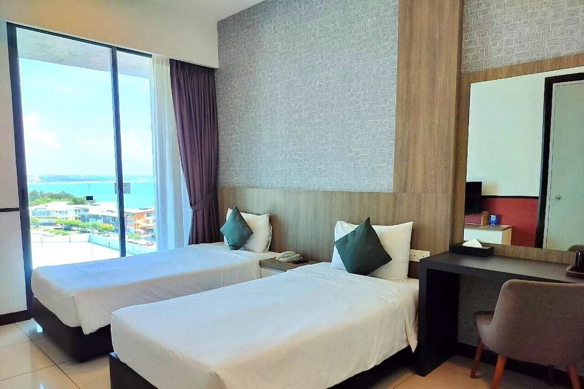 A hotel room with two beds, a desk, a chair, and a large window offering a view of the city.